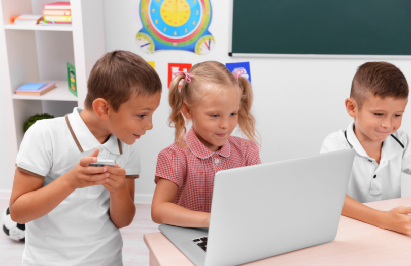 Happy primary school pupils gather round a laptop in a colourful classroom