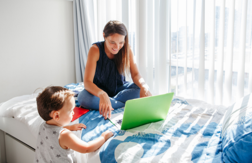 Young mum looks at laptop on a bed with toddler looking on